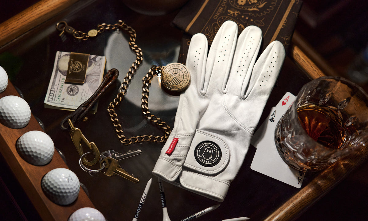 Lifestyle image of a white golf glove displayed on a counter, surrounded by keys, a ball marker chain, golf tees, a money clip, and golf balls, styled with a cool vintage aesthetic.
