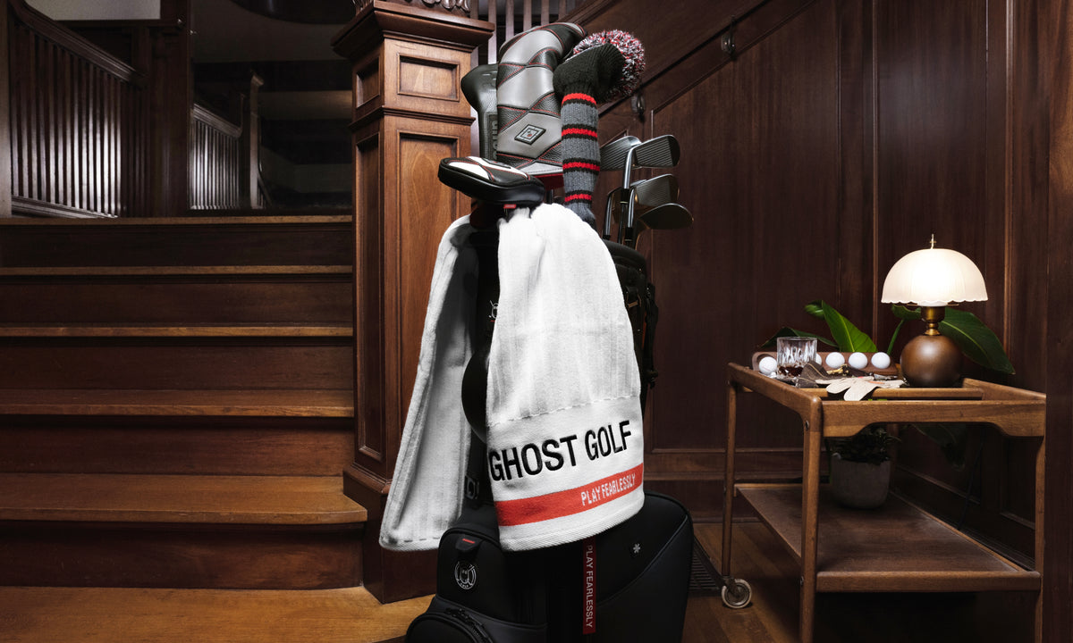 Lifestyle image of a white Ghost Golf caddie towel draped over a golf bag near a staircase in a vintage-style home, featuring Ghost Golf branding and the “Play Fearlessly” tagline.