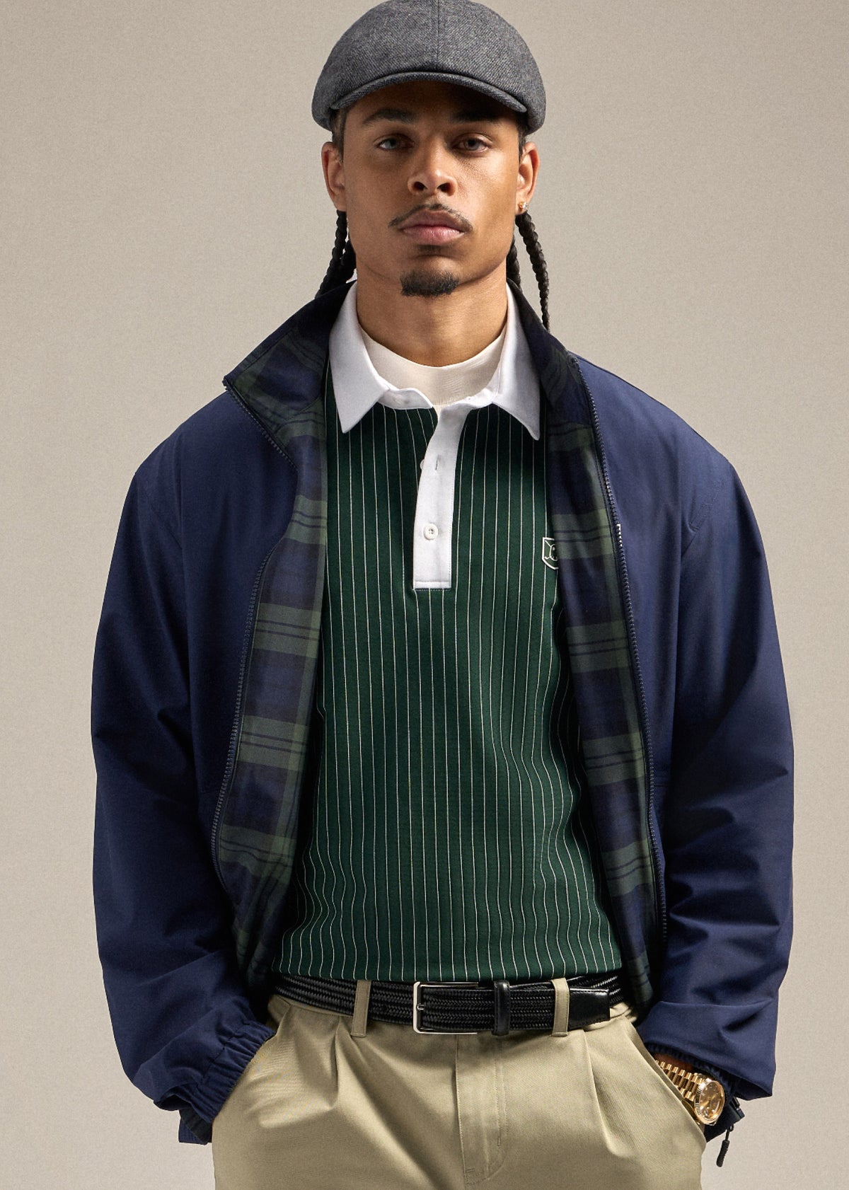 Black male model wearing a navy reversible spectator jacket with a green polo underneath. He is styled with a flat cap, sand wedge–colored shorts, and a brown belt. His hands are in his pockets, creating a relaxed and confident pose.