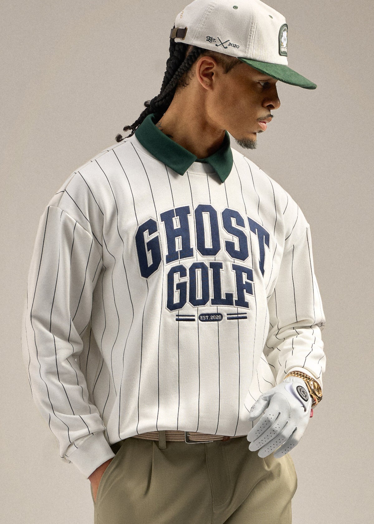 Black male model looking toward his right, wearing a white striped sweatshirt with Ghost Golf embroidered on it and a green polo underneath. He is styled with sand wedge–colored pants and a white cap, with one hand in his pocket, creating a relaxed and confident pose.