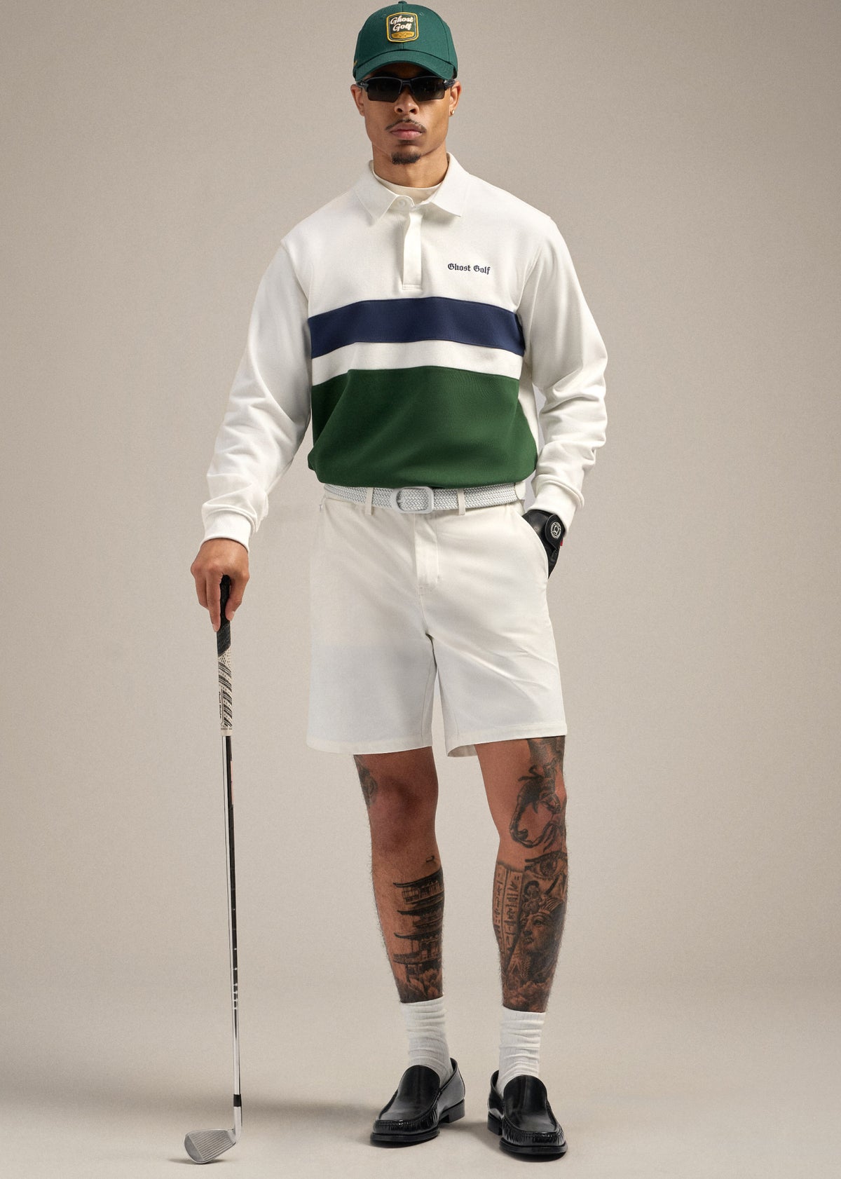 Full body shot of a Black male model wearing a white spectator polo sweatshirt paired with white shorts and a white belt. He is wearing a black glove, with his right hand in his pocket while the other hand holds a golf club resting on the floor. He is styled with a green cap, black dress shoes, and sunglasses, creating a confident and polished golf-inspired pose.