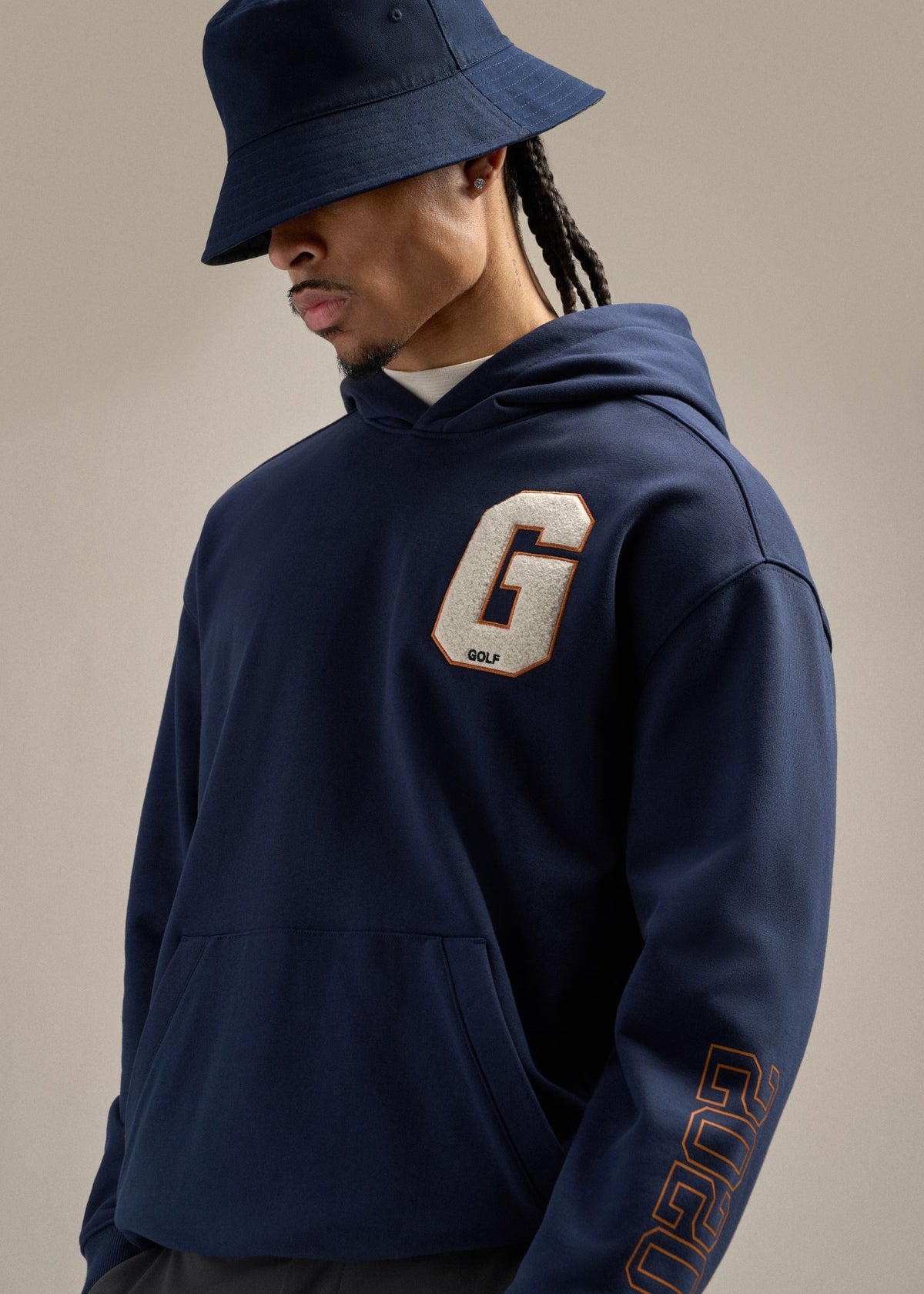 Black male model wearing a navy hoodie with a large G hit on the left chest, paired with a navy reversible bucket hat. He is looking toward his left, creating a relaxed and modern golf-inspired look.