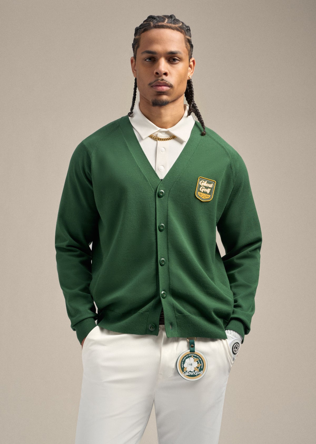 Full top shot of black male model wearing a green cardigan over a white polo. Both hands are in his pockets. He is wearing white pants with a Patrons Only golf accessory clipped to the front of the pants, creating a clean, golf-inspired look.